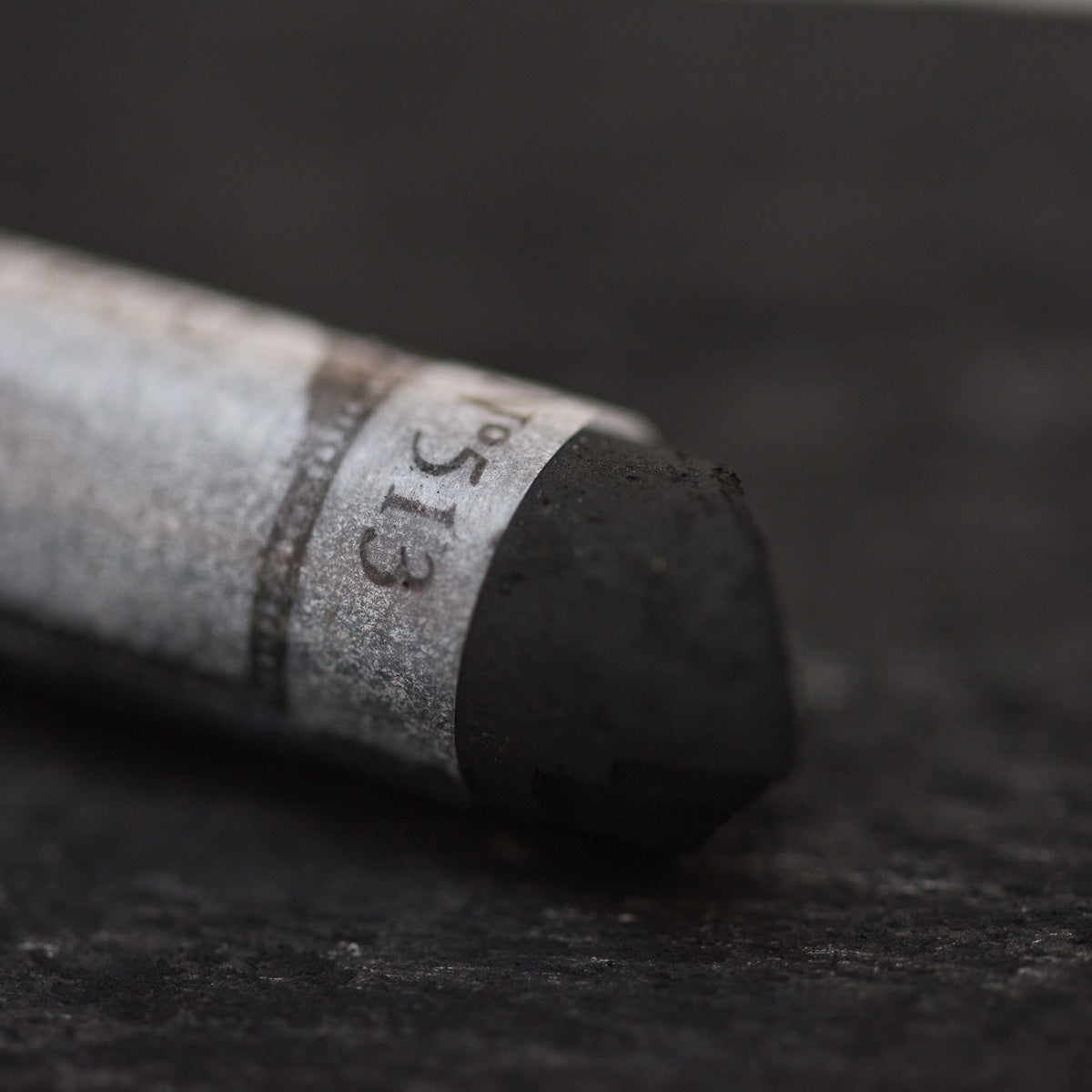 A close-up of a black Sennelier extra soft pastel. on the same coloured background, with the colour information printed on the paper wrap.