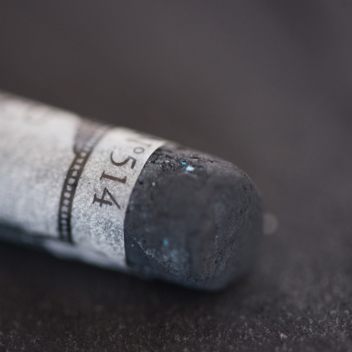 A close-up of a dark grey Sennelier extra soft pastel. on the same coloured background, with the colour information printed on the paper wrap.