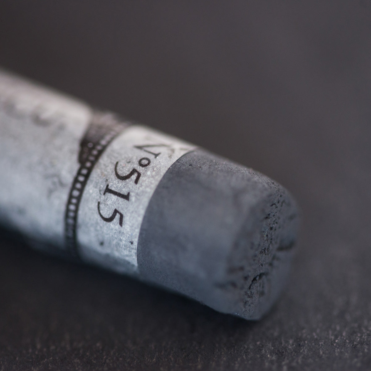A close-up of a dark grey Sennelier extra soft pastel. on the same coloured background, with the colour information printed on the paper wrap.