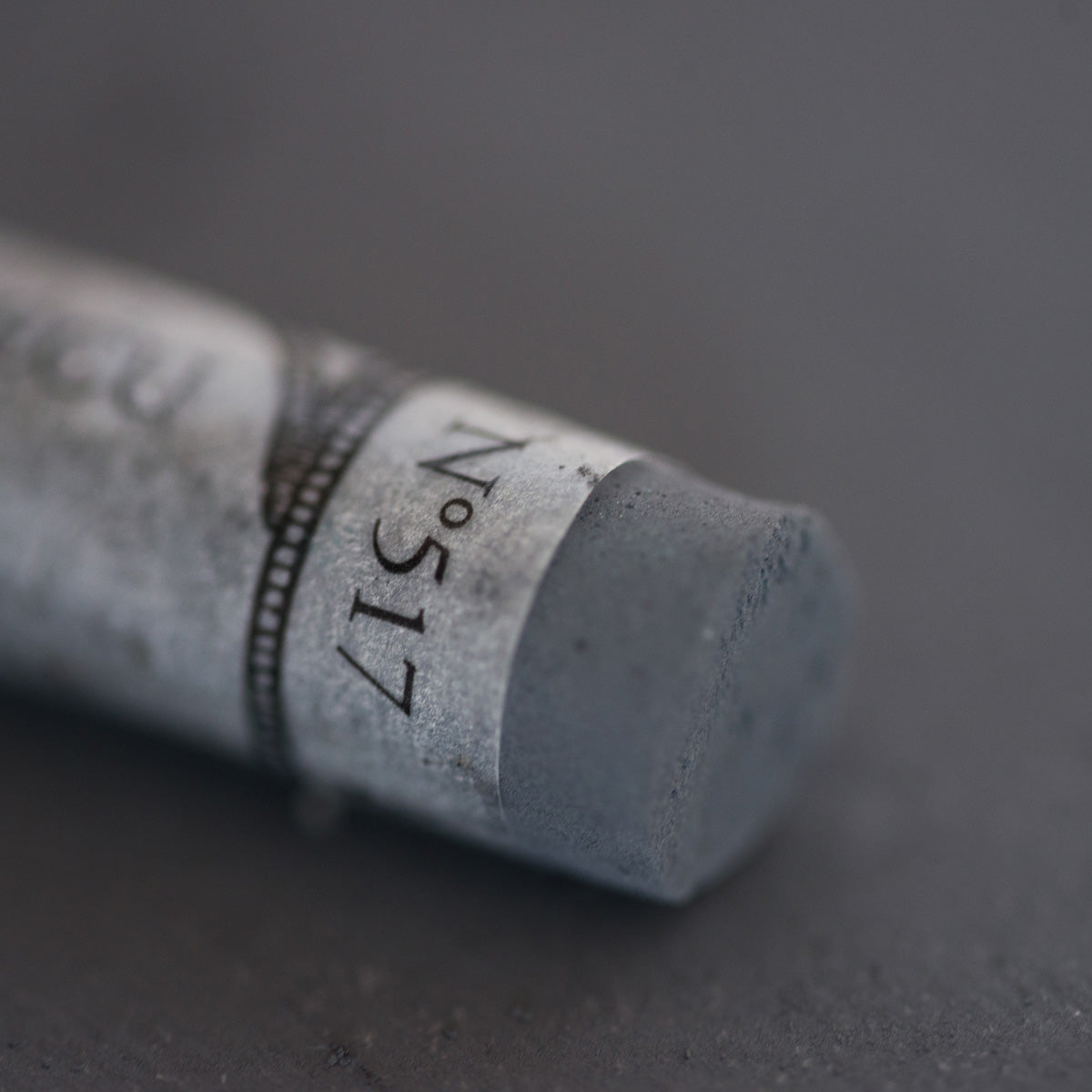 A close-up of a grey Sennelier extra soft pastel. on the same coloured background, with the colour information printed on the paper wrap.