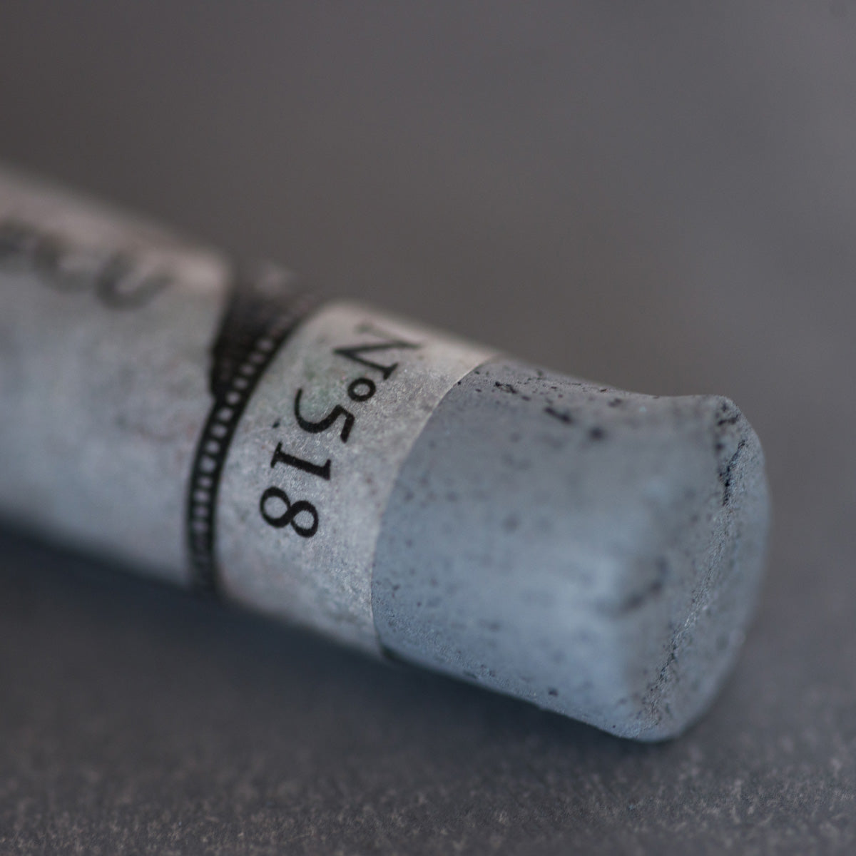 A close-up of a grey Sennelier extra soft pastel. on the same coloured background, with the colour information printed on the paper wrap.