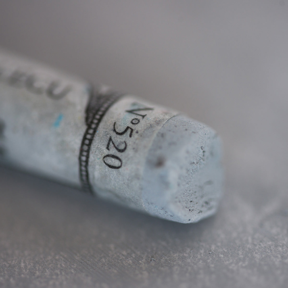 A close-up of a light grey Sennelier extra soft pastel. on the same coloured background, with the colour information printed on the paper wrap.