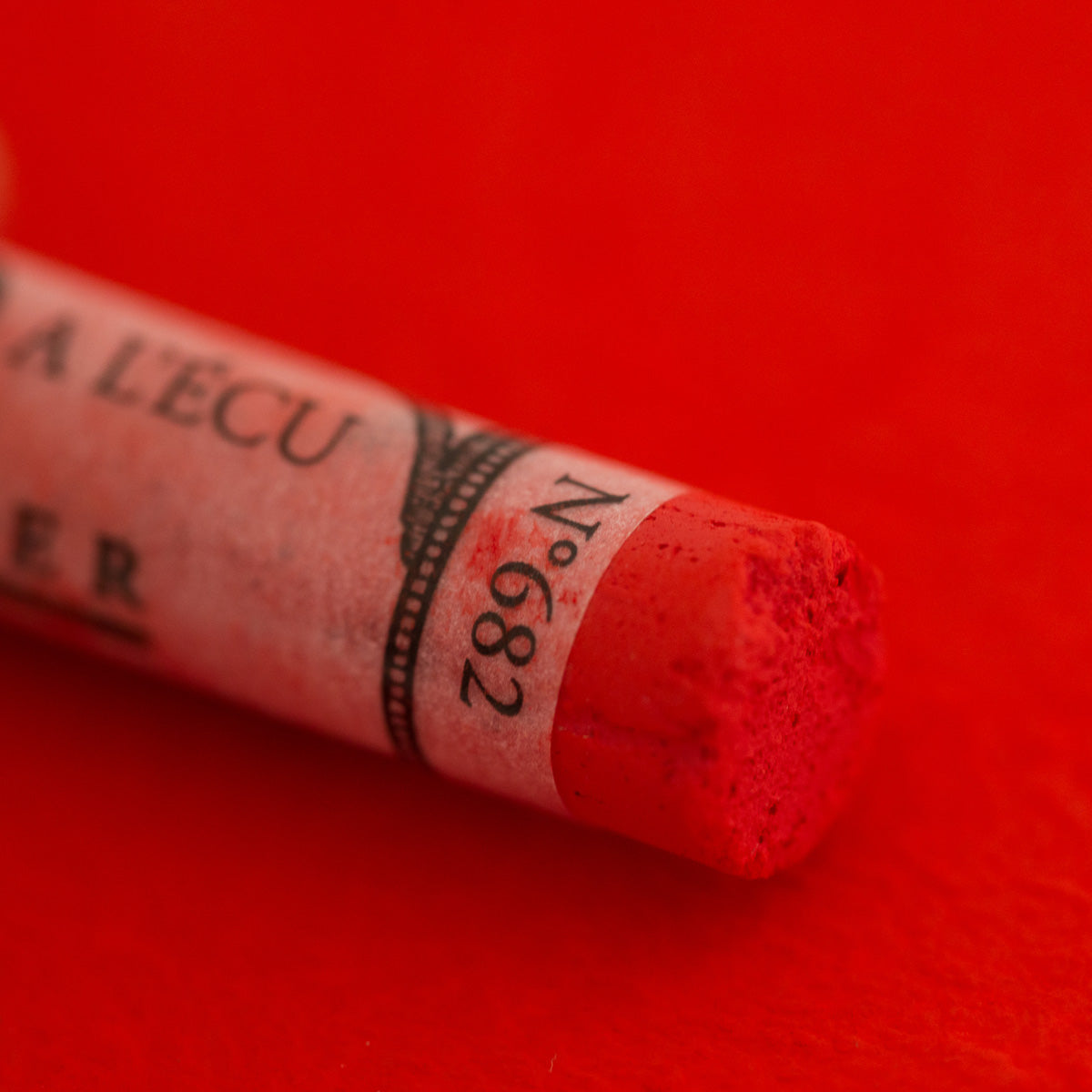 A close-up of a red Sennelier extra soft pastel on the same coloured background, with the colour information printed on the paper wrap.