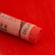 A close-up of a red Sennelier extra soft pastel on the same coloured background, with the colour information printed on the paper wrap.