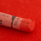 A close-up of a red Sennelier extra soft pastel on the same coloured background, with the colour information printed on the paper wrap.