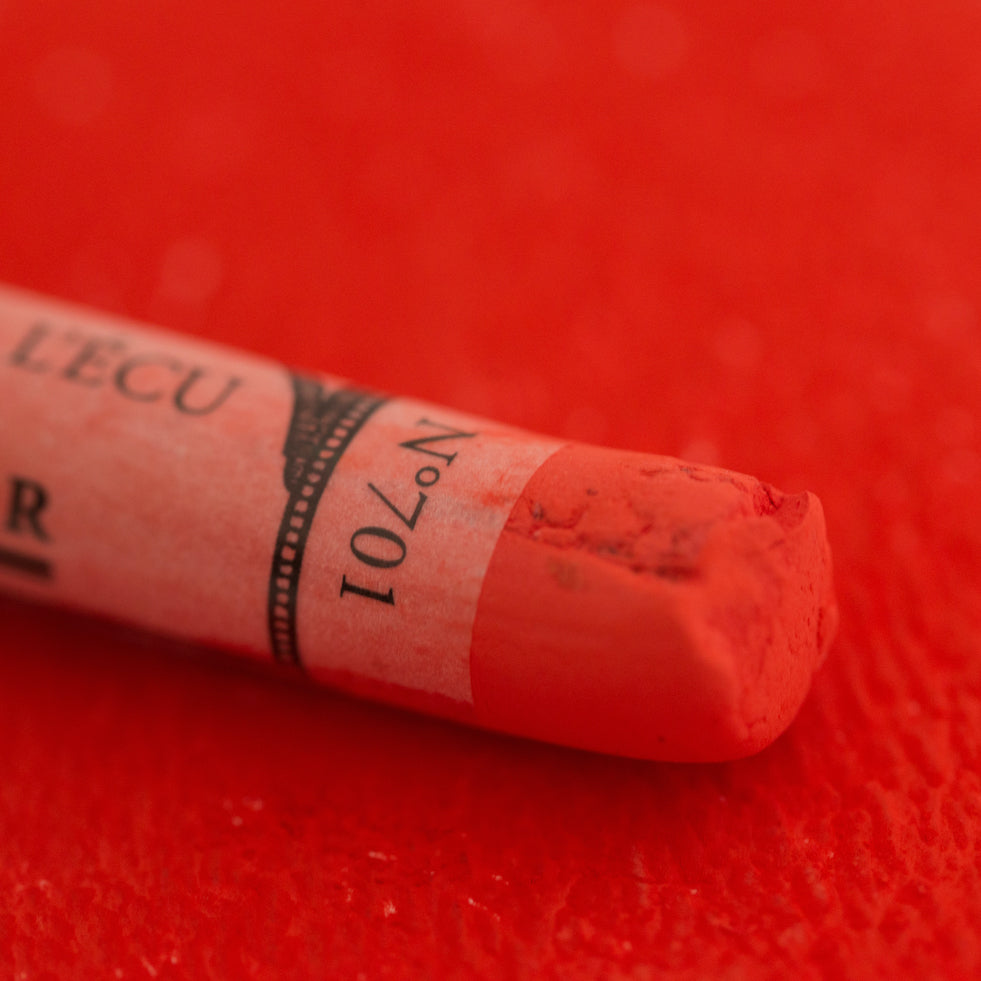 A close-up of a red Sennelier extra soft pastel on the same coloured background, with the colour information printed on the paper wrap.