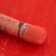A close-up of a red Sennelier extra soft pastel on the same coloured background, with the colour information printed on the paper wrap.