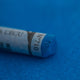 A close-up of a blue Sennelier extra soft pastel on the same coloured background, with the colour information printed on the paper wrap.