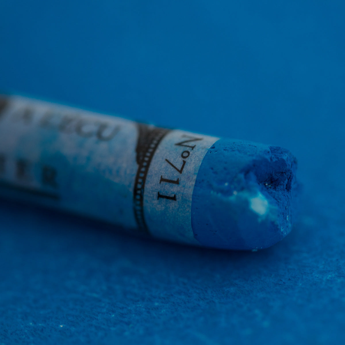 A close-up of a blue Sennelier extra soft pastel on the same coloured background, with the colour information printed on the paper wrap.