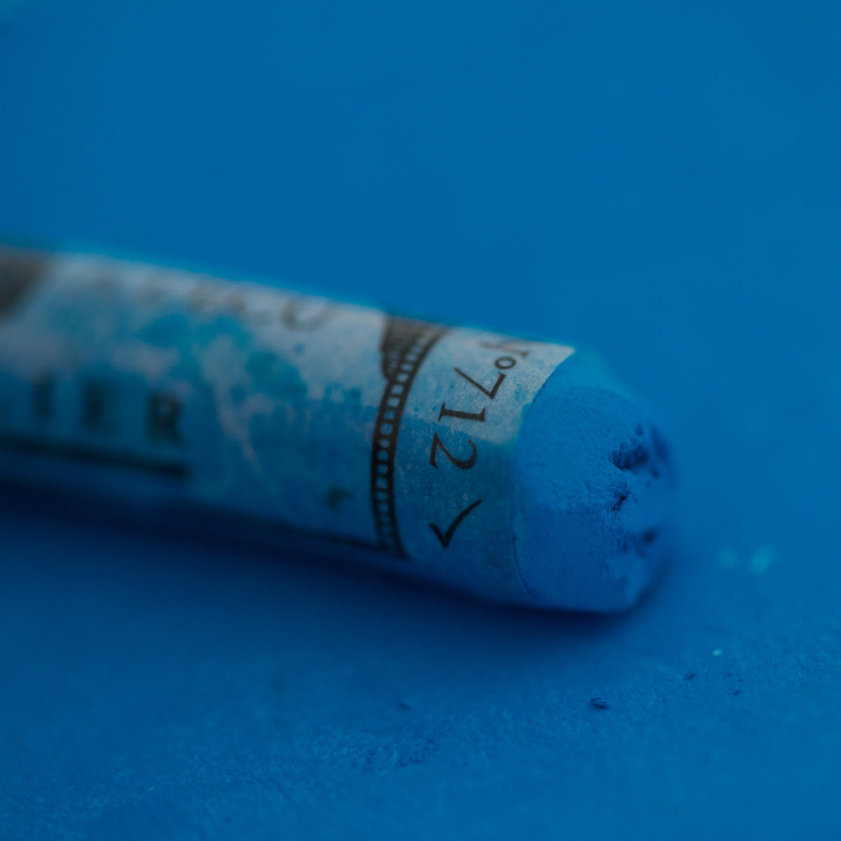 A close-up of a blue Sennelier extra soft pastel on the same coloured background, with the colour information printed on the paper wrap.