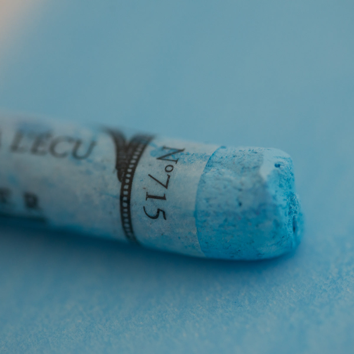 A close-up of a blue Sennelier extra soft pastel on the same coloured background, with the colour information printed on the paper wrap.