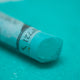 A close-up of a green Sennelier extra soft pastel. on the same coloured background, with the colour information printed on the paper wrap.