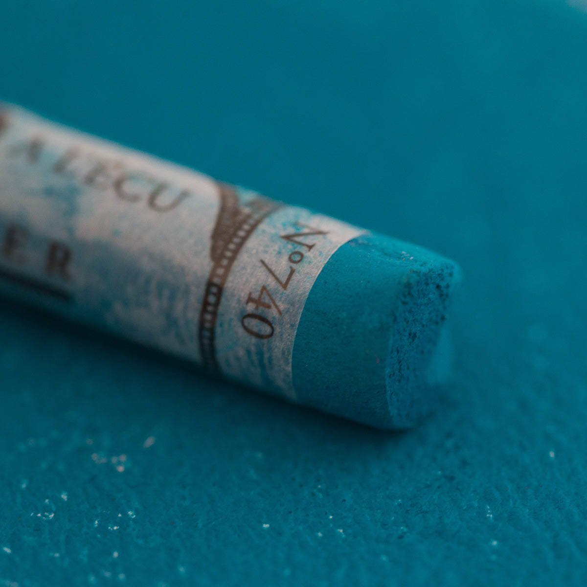 A close-up of a blue/green Sennelier extra soft pastel. on the same coloured background, with the colour information printed on the paper wrap.
