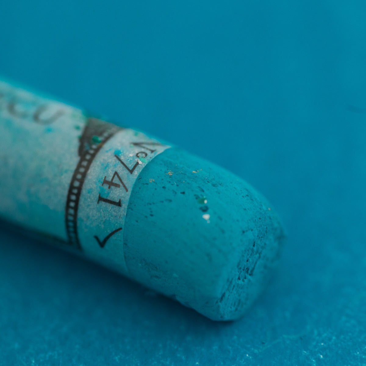 A close-up of a blue/green Sennelier extra soft pastel. on the same coloured background, with the colour information printed on the paper wrap.