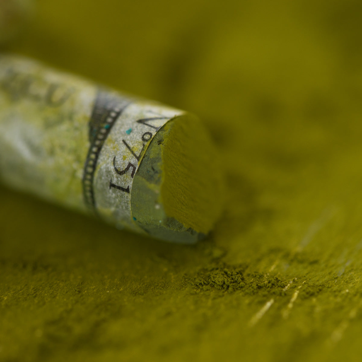 A close-up of a green Sennelier extra soft pastel, on the same coloured background, with the colour information printed on the paper wrap.