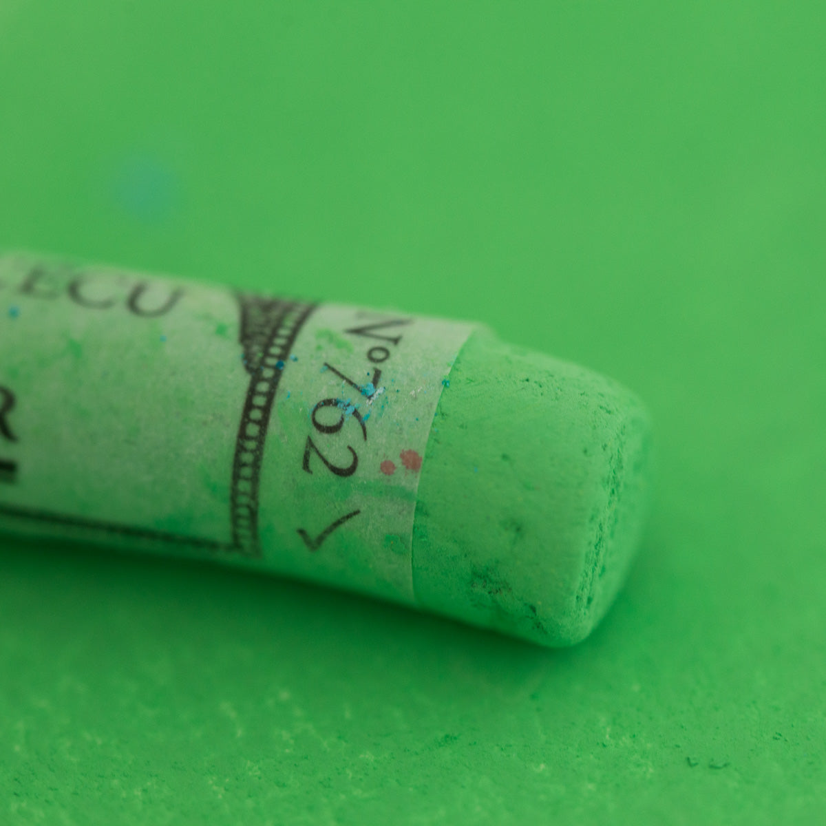 A close-up of a bright green Sennelier extra soft pastel. on the same coloured background, with the colour information printed on the paper wrap.