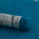 A close-up of a blue Sennelier extra soft pastel on the same coloured background, with the colour information printed on the paper wrap.
