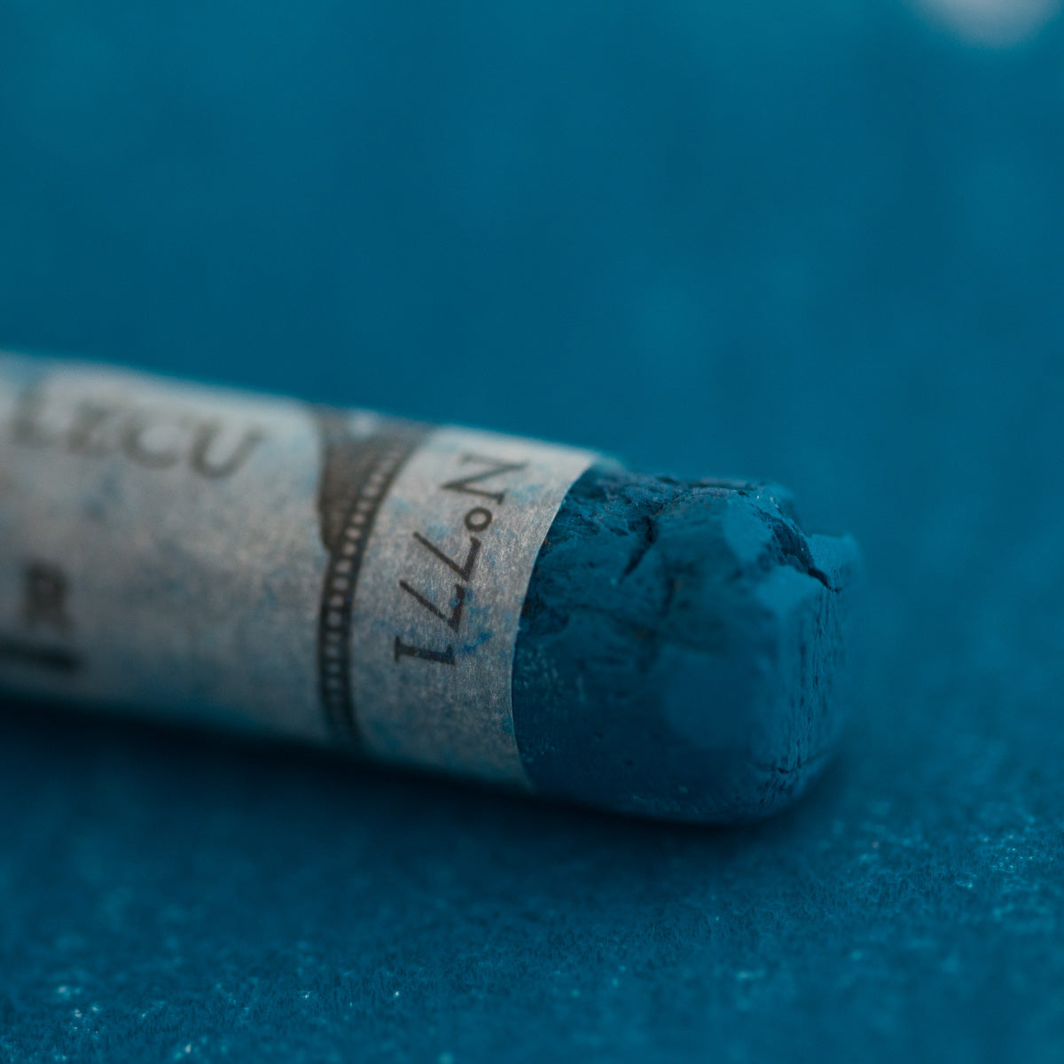 A close-up of a blue Sennelier extra soft pastel on the same coloured background, with the colour information printed on the paper wrap.