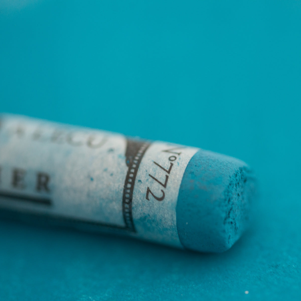 A close-up of a blue Sennelier extra soft pastel on the same coloured background, with the colour information printed on the paper wrap.