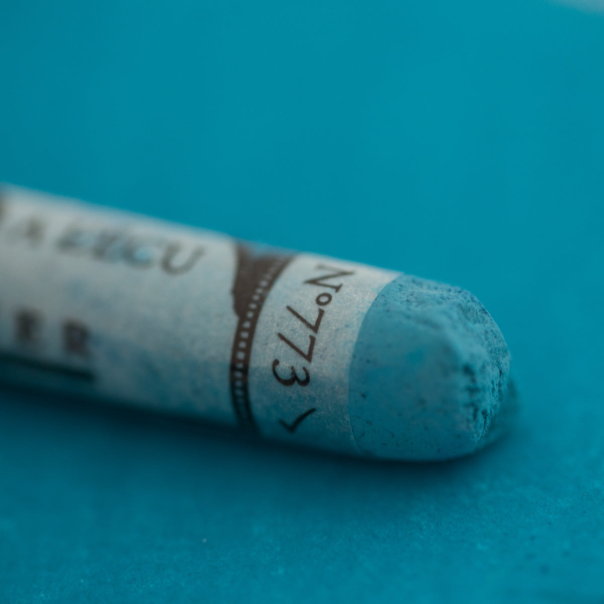 A close-up of a blue Sennelier extra soft pastel on the same coloured background, with the colour information printed on the paper wrap.