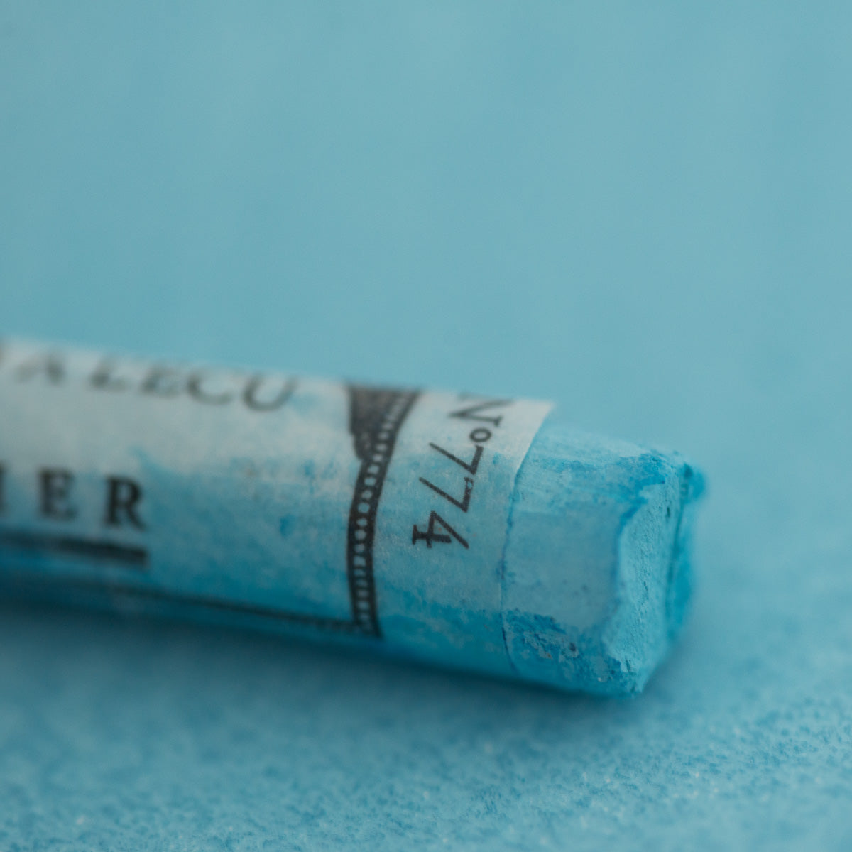 A close-up of a blue Sennelier extra soft pastel on the same coloured background, with the colour information printed on the paper wrap.