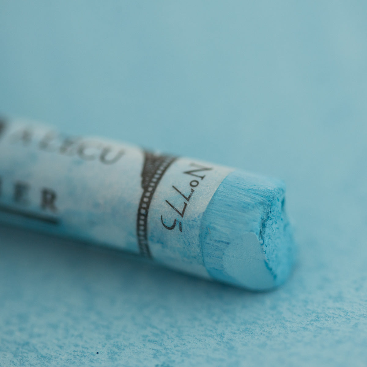 A close-up of a light blue Sennelier extra soft pastel on the same coloured background, with the colour information printed on the paper wrap.
