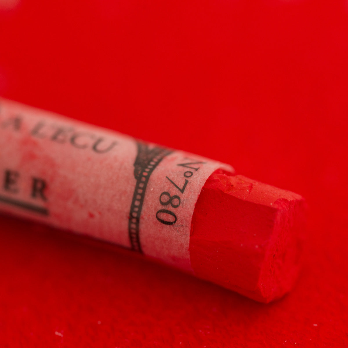 A close-up of a bright red Sennelier extra soft pastel on the same coloured background, with the colour information printed on the paper wrap.