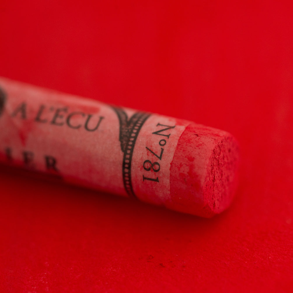A close-up of a bright red Sennelier extra soft pastel on the same coloured background, with the colour information printed on the paper wrap.