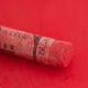 A close-up of a red Sennelier extra soft pastel on the same coloured background, with the colour information printed on the paper wrap.