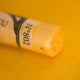 A close-up of an iridescent yellow Sennelier extra soft pastel. on the same coloured background, with the colour information printed on the paper wrap.