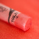 A close-up of an iridescent red Sennelier extra soft pastel. on the same coloured background, with the colour information printed on the paper wrap.