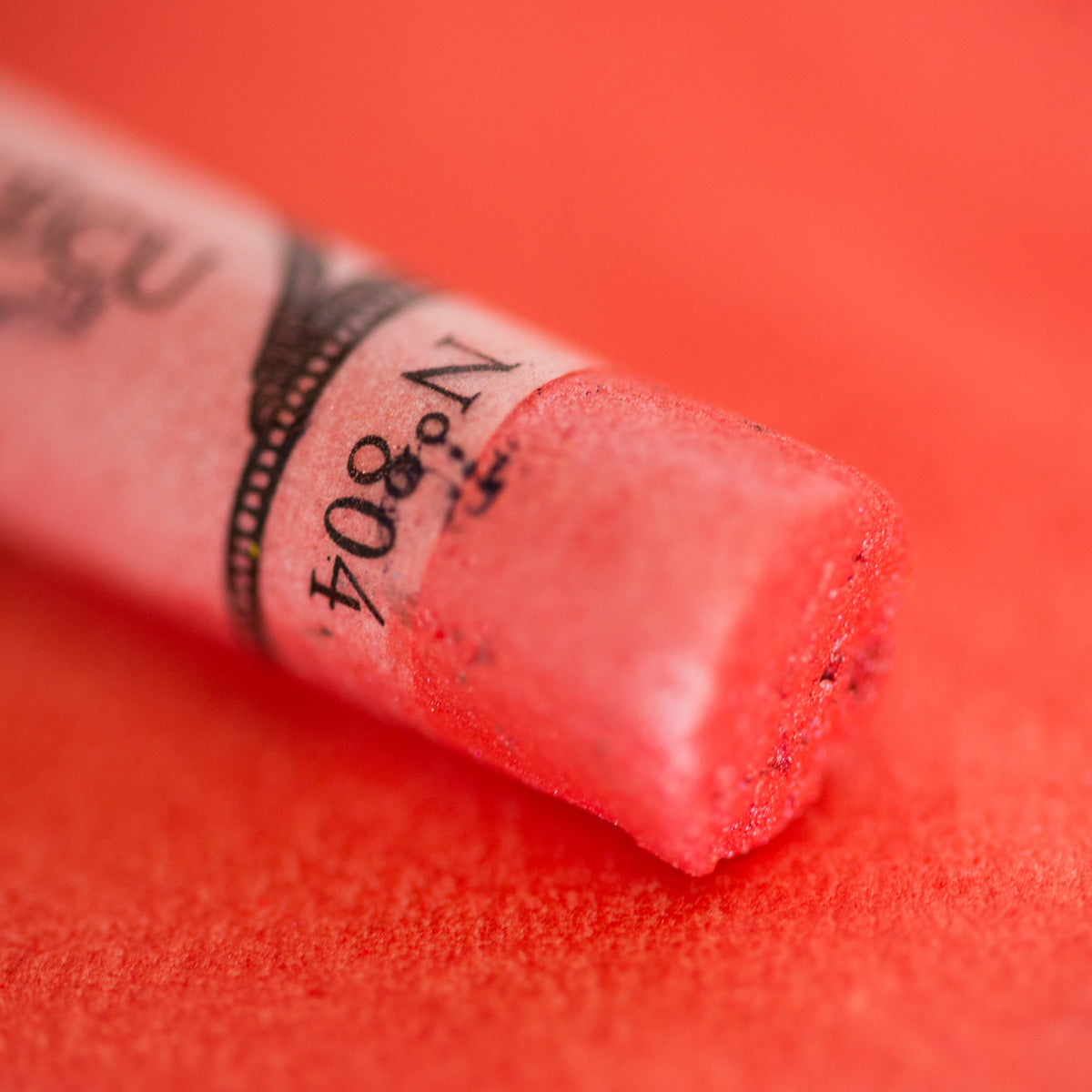 A close-up of an iridescent red Sennelier extra soft pastel. on the same coloured background, with the colour information printed on the paper wrap.