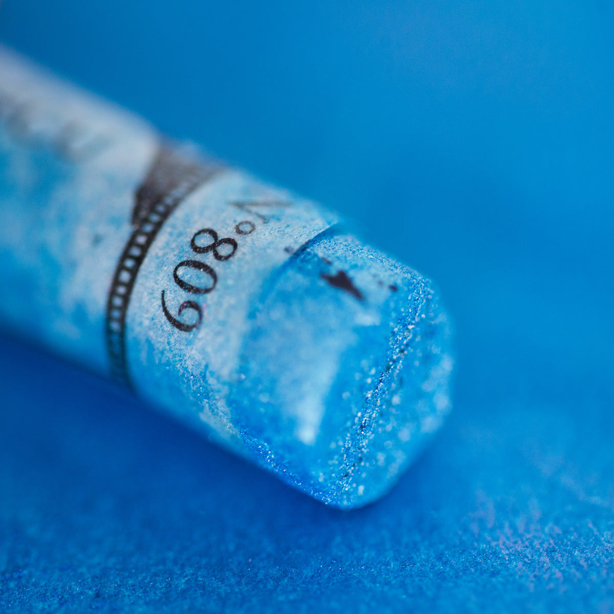 A close-up of an iridescent blue Sennelier extra soft pastel. on the same coloured background, with the colour information printed on the paper wrap.