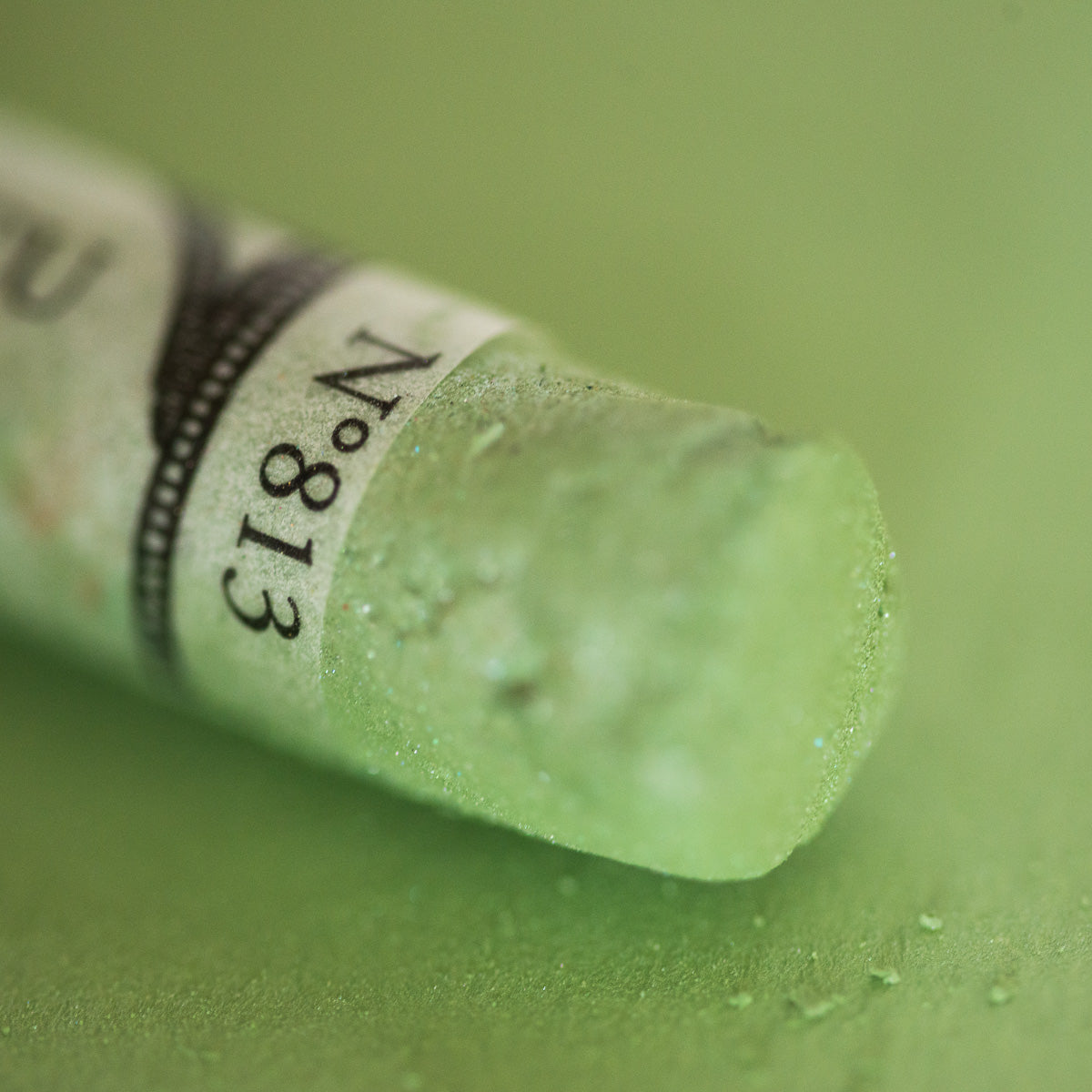 A close-up of an iridescent green Sennelier extra soft pastel. on the same coloured background, with the colour information printed on the paper wrap.