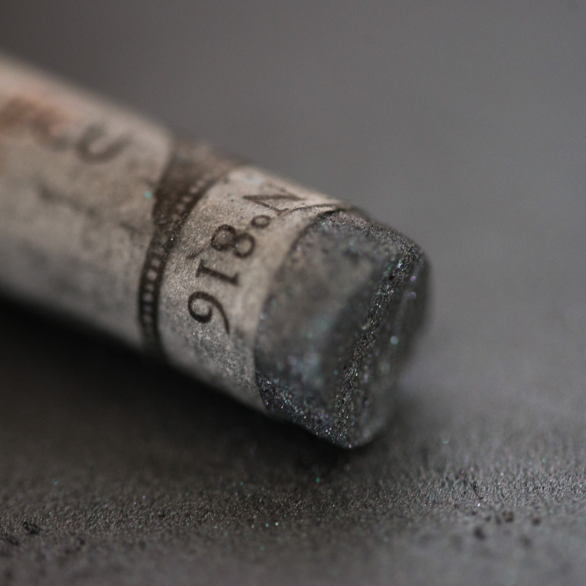 A close-up of a black iridescent Sennelier extra soft pastel. on the same coloured background, with the colour information printed on the paper wrap.