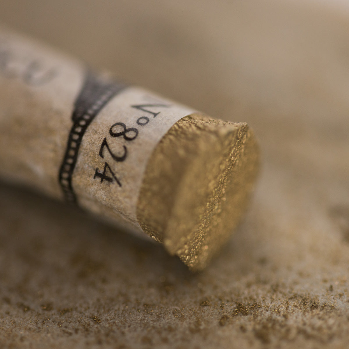 A close-up of an iridescent gold Sennelier extra soft pastel. on the same coloured background, with the colour information printed on the paper wrap.