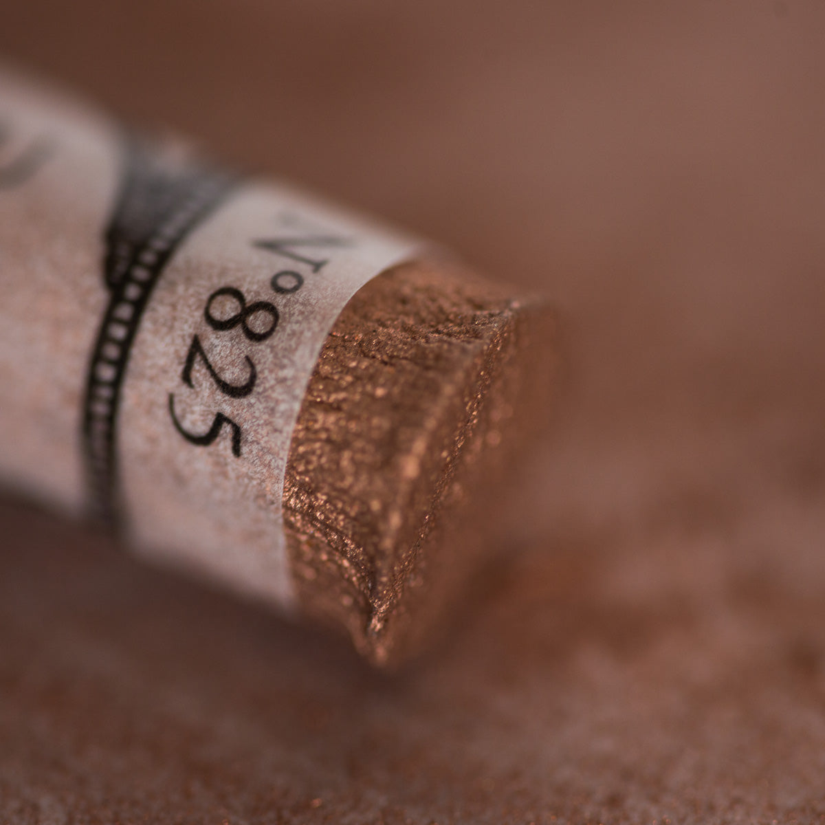 A close-up of an iridescent copper Sennelier extra soft pastel. on the same coloured background, with the colour information printed on the paper wrap.