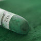 A close-up of a green Sennelier extra soft pastel, on the same coloured background, with the colour information printed on the paper wrap.