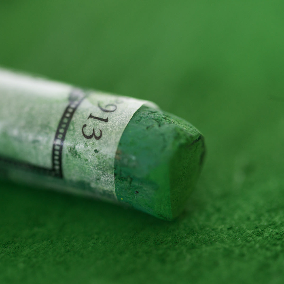 A close-up of a green Sennelier extra soft pastel, on the same coloured background, with the colour information printed on the paper wrap.