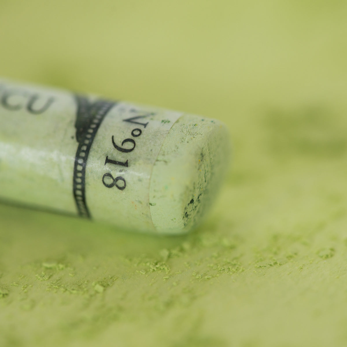 A close-up of a light green Sennelier extra soft pastel, on the same coloured background, with the colour information printed on the paper wrap.
