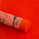 A close-up of an orange Sennelier extra soft pastel on the same coloured background, with the colour information printed on the paper wrap.