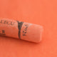 A close-up of a light orange Sennelier extra soft pastel on the same coloured background, with the colour information printed on the paper wrap.
