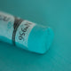 A close-up of a green Sennelier extra soft pastel. on the same coloured background, with the colour information printed on the paper wrap.