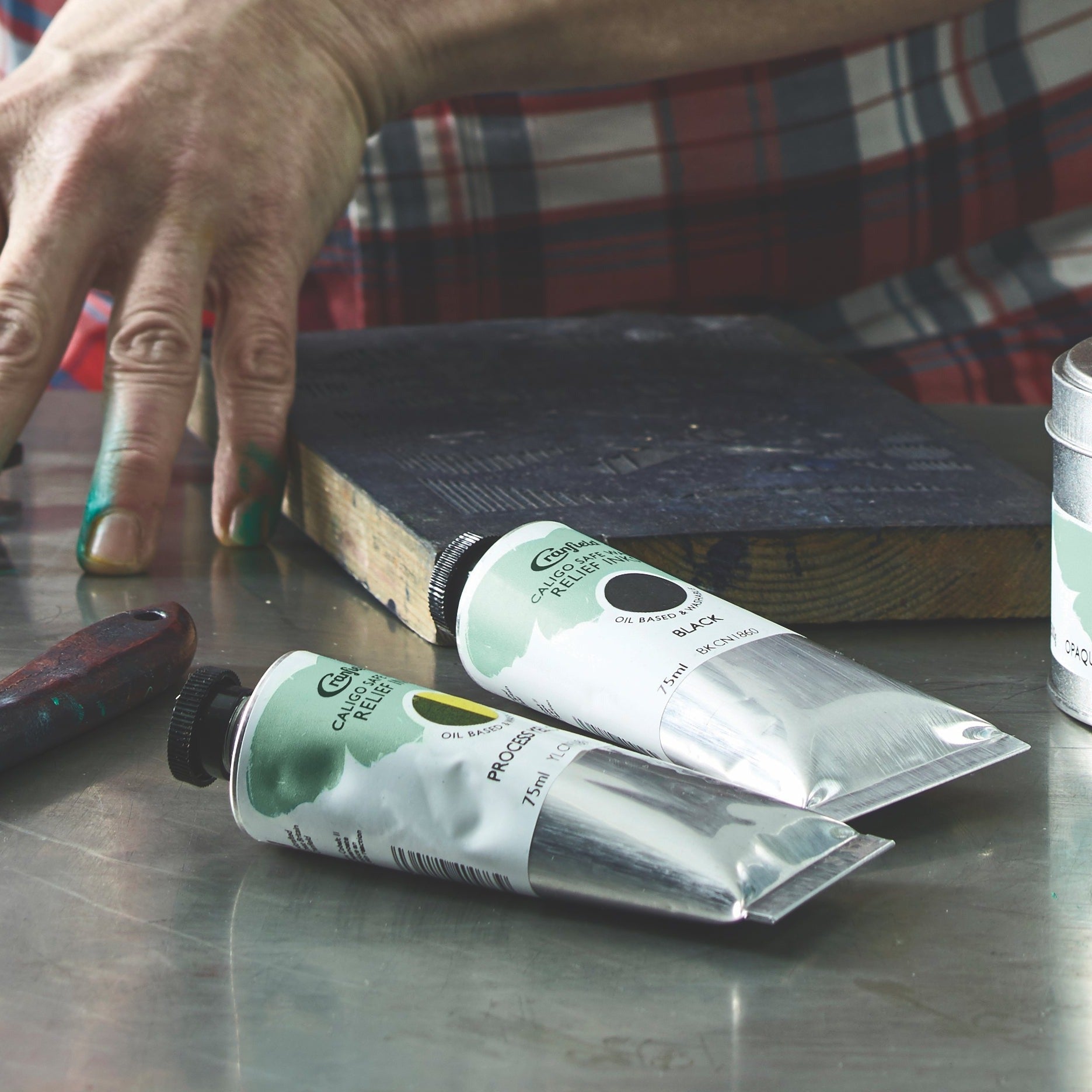 A person's hand reaching for a tube of Cranfield Caligo Relief Ink, with other tubes and a varnish brush on a surface, suggesting an artistic environment.