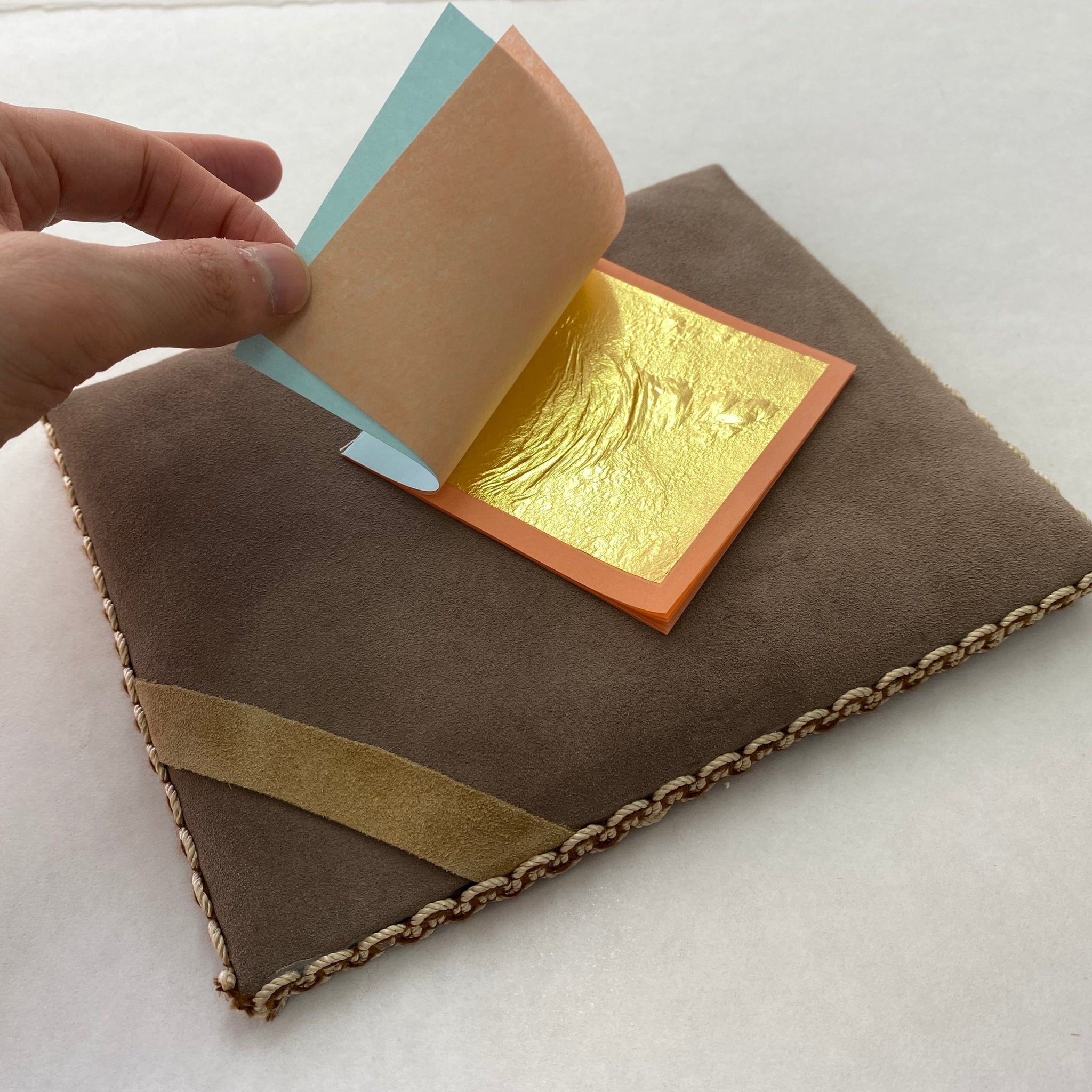 A person's hand lifting a corner of a paper stack revealing a  sheet of 23 carat gold leaf underneath. Sitting on a suede gilding cushion.
