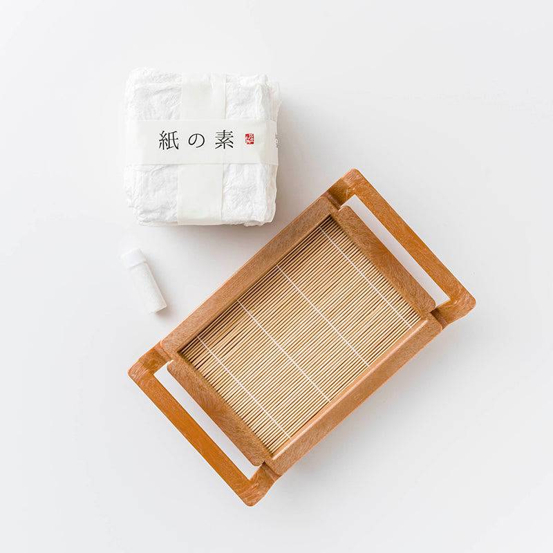A Japanese papermaking frame made of plastic with a bamboo mat, a set of compressed pulp sheets, and a bottle of binding agent displayed on a white background.
